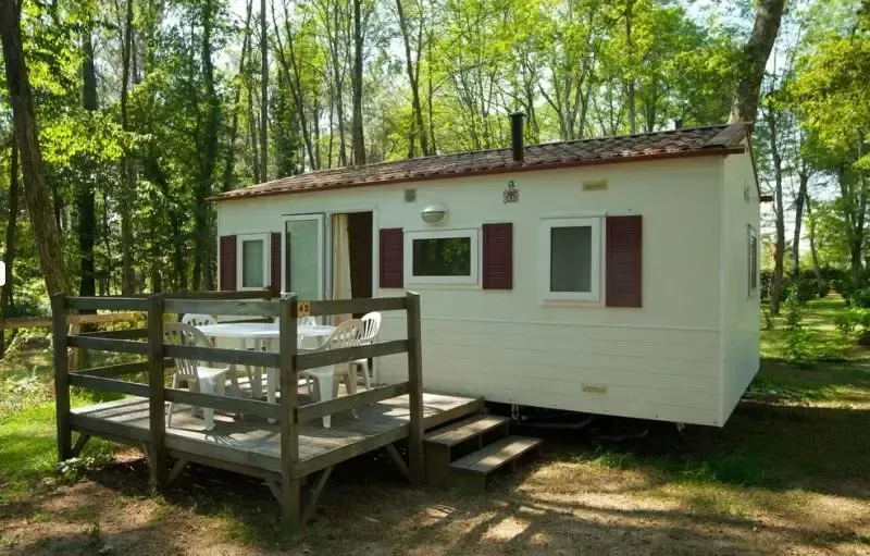 Mobil-home blanc avec volets bordeaux et terrasse en bois dans un environnement arboré