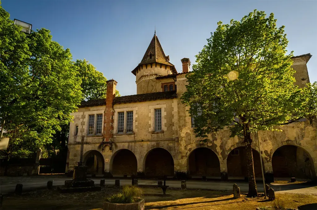 Bâtiment ancien en pierre avec tour et galerie d’arcades au milieu de grands arbres